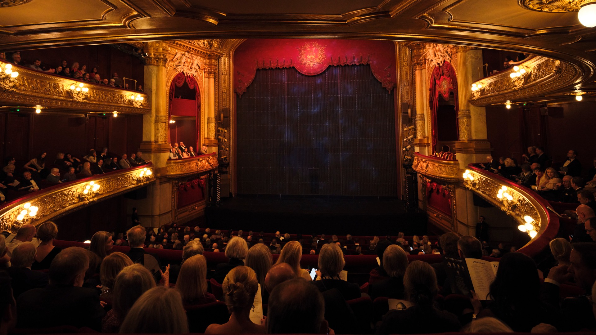 Vy från balkong mot en scen, fullt med publik och salongen är rikt ornamenterad.