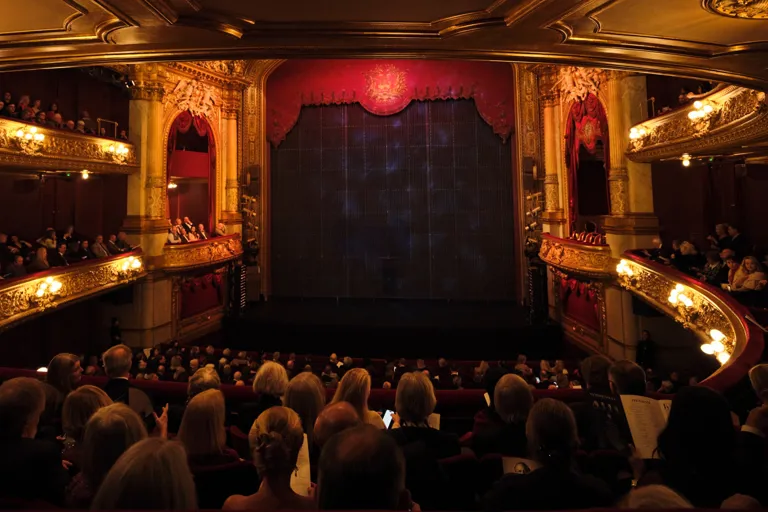 Vy från balkong mot en scen, fullt med publik och salongen är rikt ornamenterad.