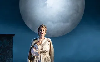 En kvinna i mantel och med krona på huvudet, i bakgrunden en stor måne.