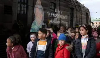 A group of children of various ages wait in front of a bus on the sidewalk.