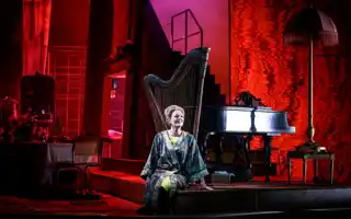 A woman sits in a room lit in red, behind her a harp and a piano.