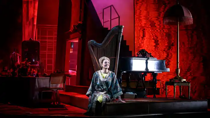 A woman sits in a room lit in red, behind her a harp and a piano.