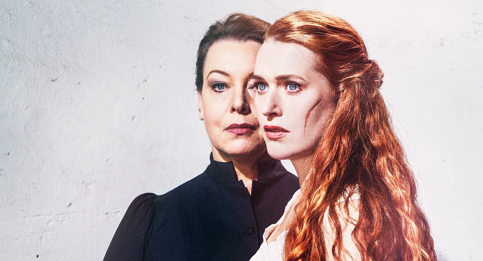 Two women against a light background, in the foreground a red-haired woman with a scar on her cheek.