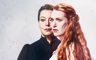 Two women against a light background, in the foreground a red-haired woman with a scar on her cheek.