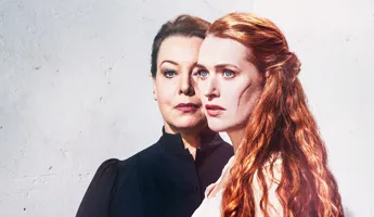 Två kvinnor mot en ljus bakgrund, i förgrunden en rödhårig kvinna med ärr på kinden.