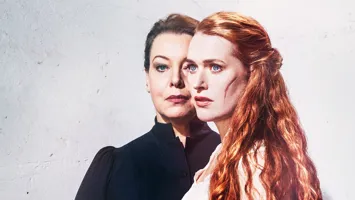 Two women against a light background, in the foreground a red-haired woman with a scar on her cheek.