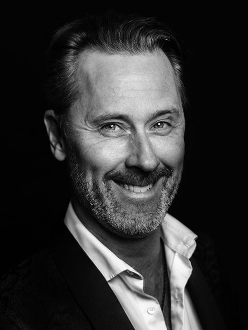 A close-up of a smiling man, he is wearing a dark suit and has a light unbuttoned shirt.