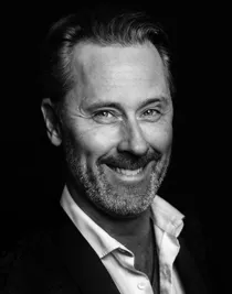 En närbild på en leende man, han bär mörk kostym och har en ljus uppknäppt skjorta.