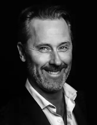 En närbild på en leende man, han bär mörk kostym och har en ljus uppknäppt skjorta.