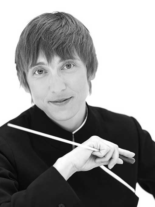 A close-up of a short-haired woman holding a conductor's baton in her hand.