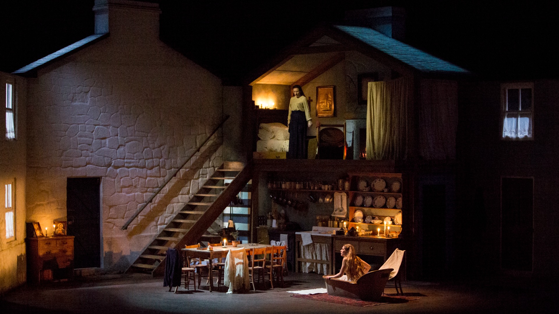 A scene with a large house that resembles a dollhouse: on the upper floor a woman stands, on the ground floor a woman sits in a bathtub.