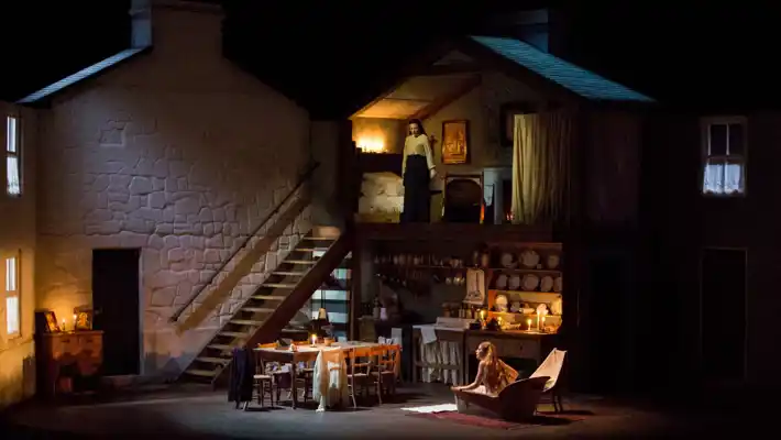 A scene with a large house that resembles a dollhouse: on the upper floor a woman stands, on the ground floor a woman sits in a bathtub.