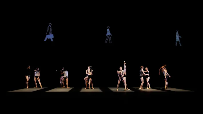 Twelve dancers on a stage, three projections of people are visible in the background.