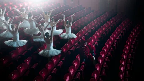 En grupp ballerinor springer mellan rader av röda stolar.