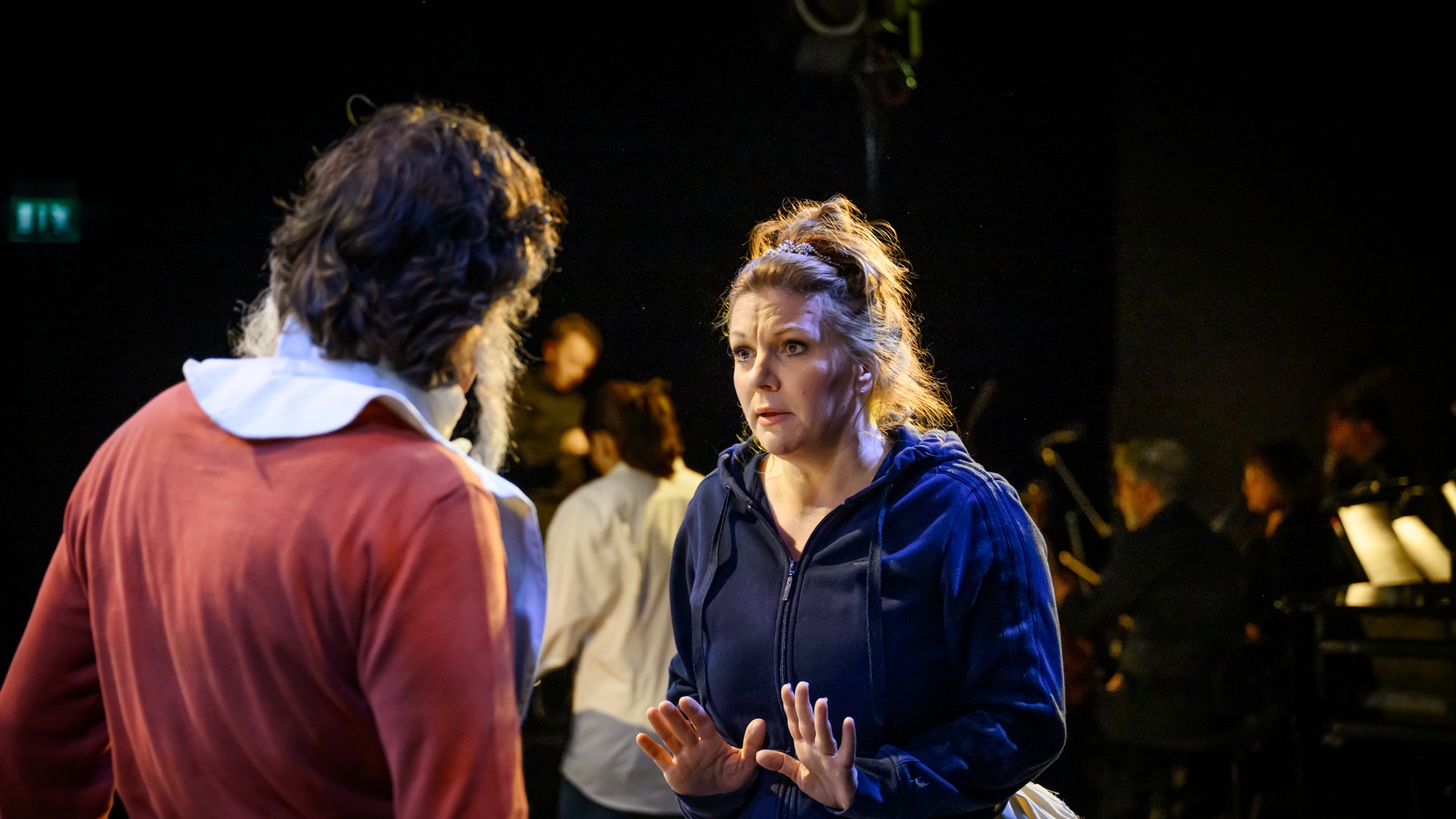 A man in red stands with his back turned to us, a woman in a blue hoodie appears to be talking to him.