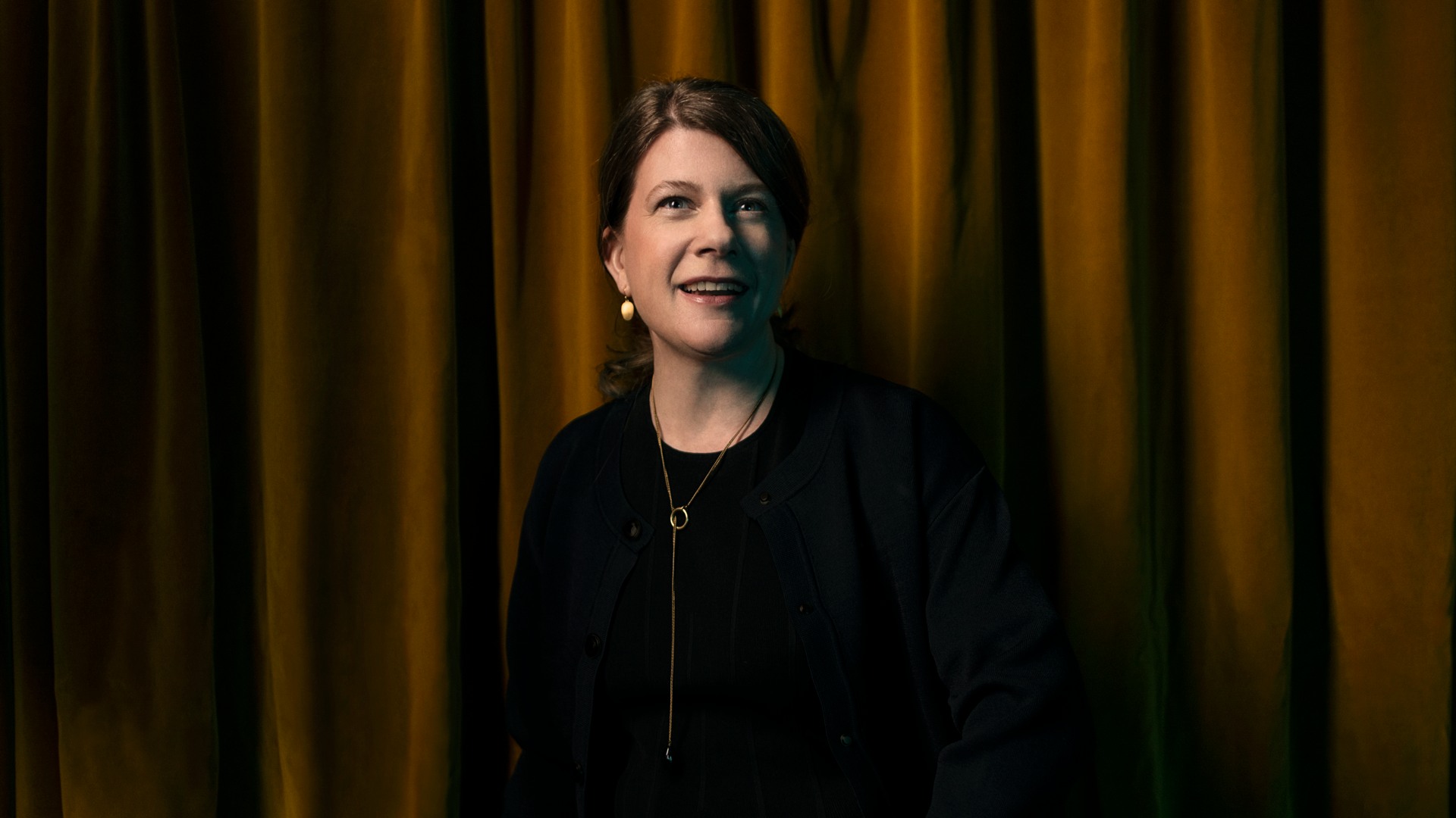 A woman with a necklace and earrings stands against a velvet curtain.