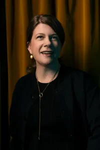 En kvinna med halsband och örhängen står mot ett sammetsdraperi.