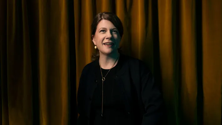 En kvinna med halsband och örhängen står mot ett sammetsdraperi.