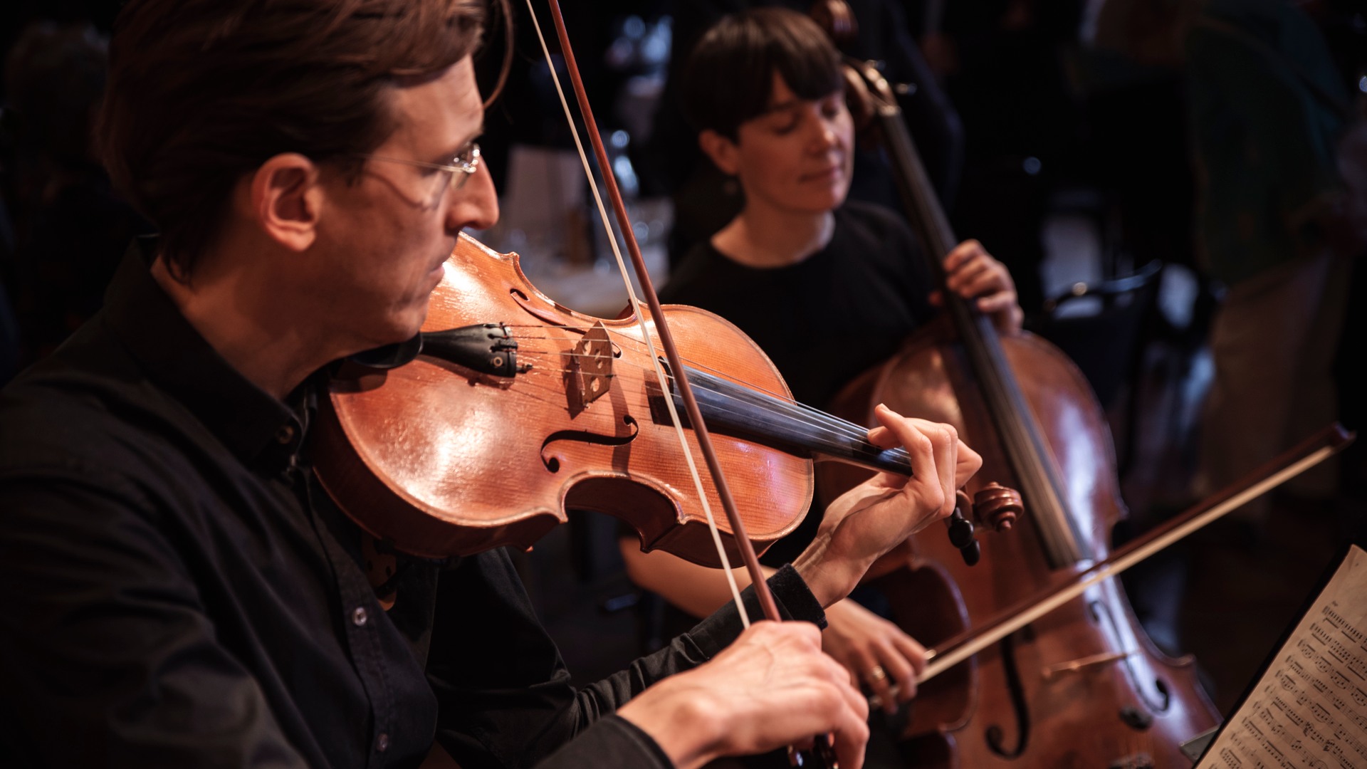 A man plays a viola in the foreground, in the background a woman plays a cello.