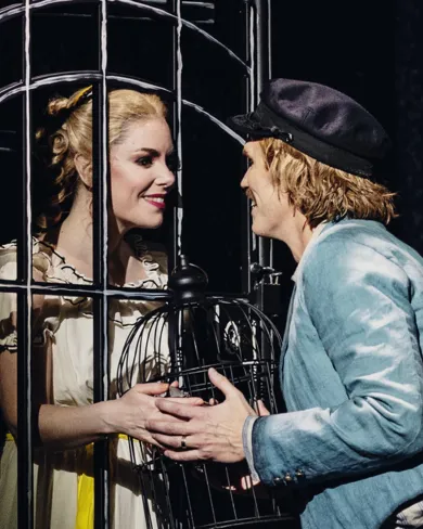 A woman stands behind a grate, a man on the other side, they hold a cage.