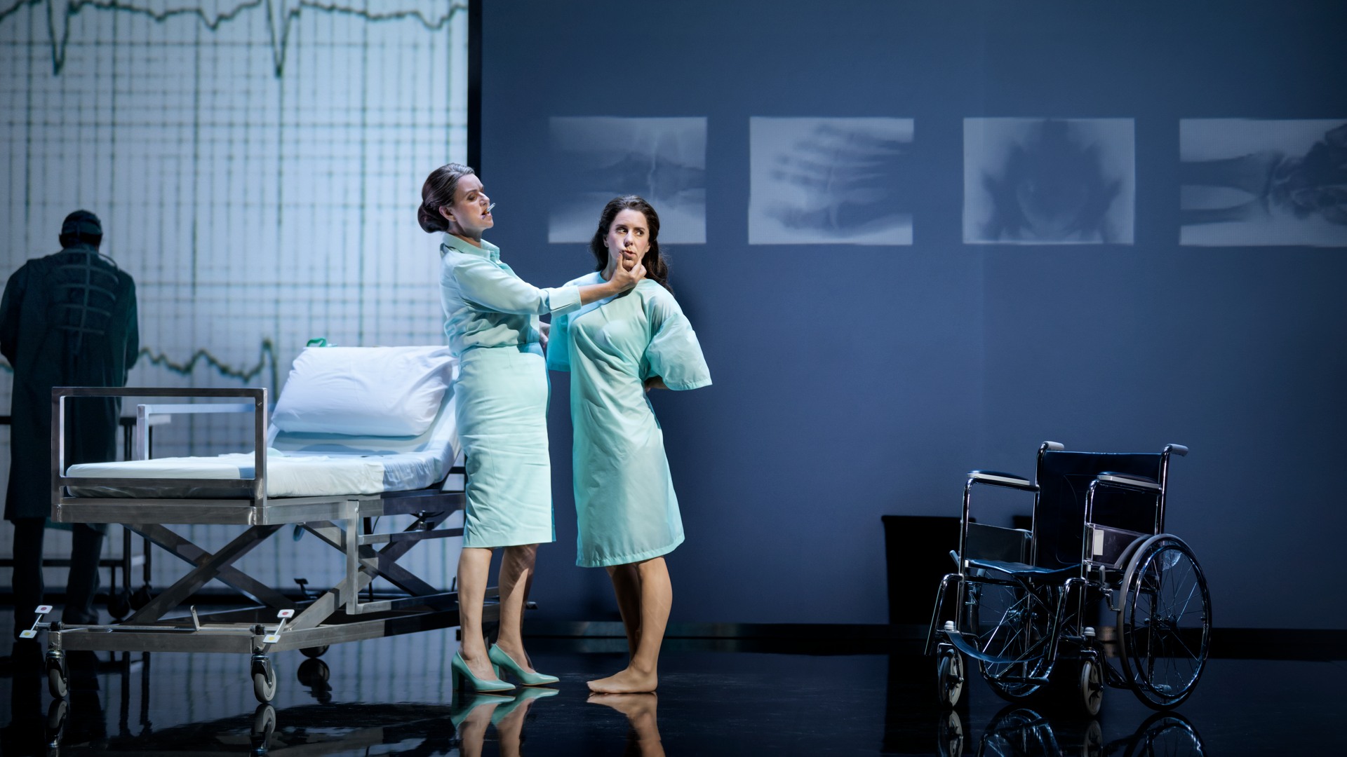 Två kvinnor står i ett sjukhusliknande rum: de är klädda i sjukhuskläder och bakom en säng och en rullstol.
