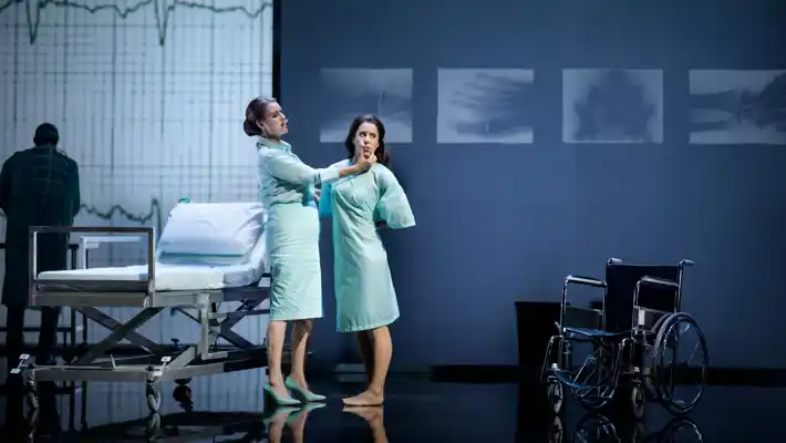 Två kvinnor står i ett sjukhusliknande rum: de är klädda i sjukhuskläder och bakom en säng och en rullstol.