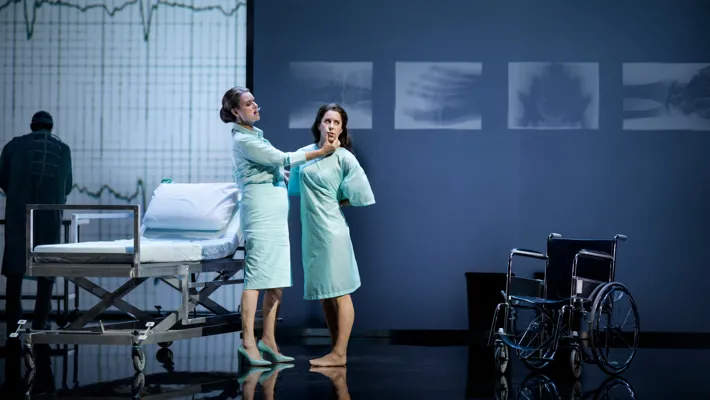 Two women are standing in a hospital-like room: they are dressed in hospital clothes and behind a bed and a wheelchair.