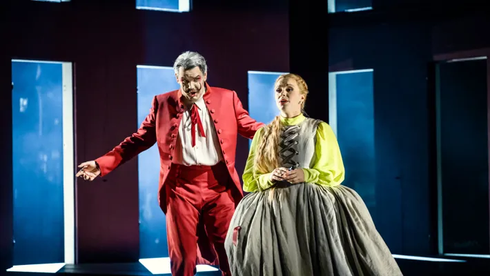 En rödklädd man och en kvinna i stor klänning står i ett rum med flera öppningar.