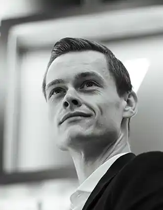 En närbild på en man, tagen lätt underifrån: han bär kostym och blickar uppåt, utåt.