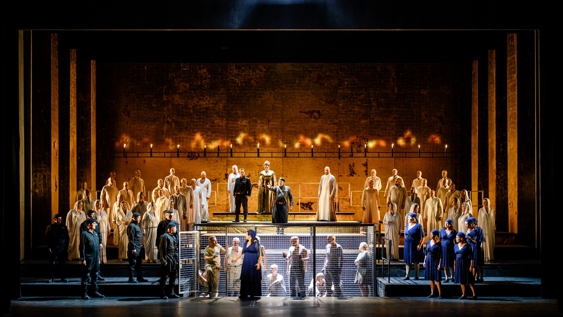 En guldprydd scen med olika personer ståendes på gradänger, i bakgrunden eld, i förgrunden en bur.