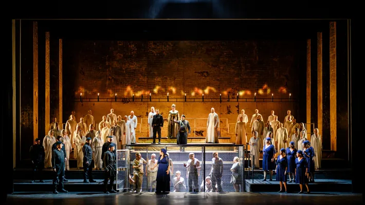 En guldprydd scen med olika personer ståendes på gradänger, i bakgrunden eld, i förgrunden en bur.