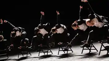 Six dancers dressed in black and white costumes jump over chairs.