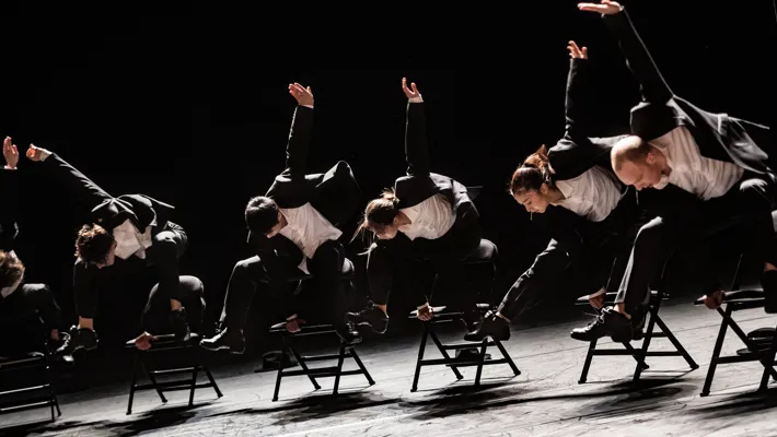 Sex dansare iklädda svart-vita kostymer hoppar över stolar.