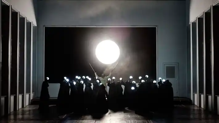 I en stor sal står många bemantlade personer med ljusbollar i händerna, längst fram lyfter en man sina kryckor mot en stor måne i fonden.