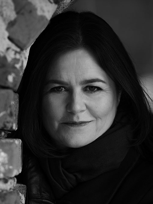 A close-up of a woman in dark clothes, standing partly behind a wall on the left side.