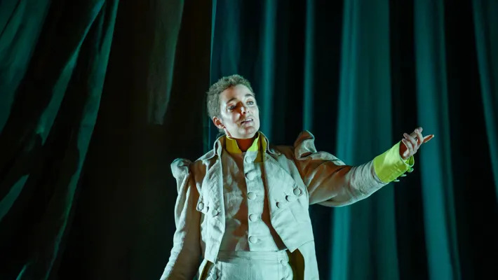 En ung man i kostym står mot ett mörkgrönt tygstycke och sträcker ut vänsterhanden.