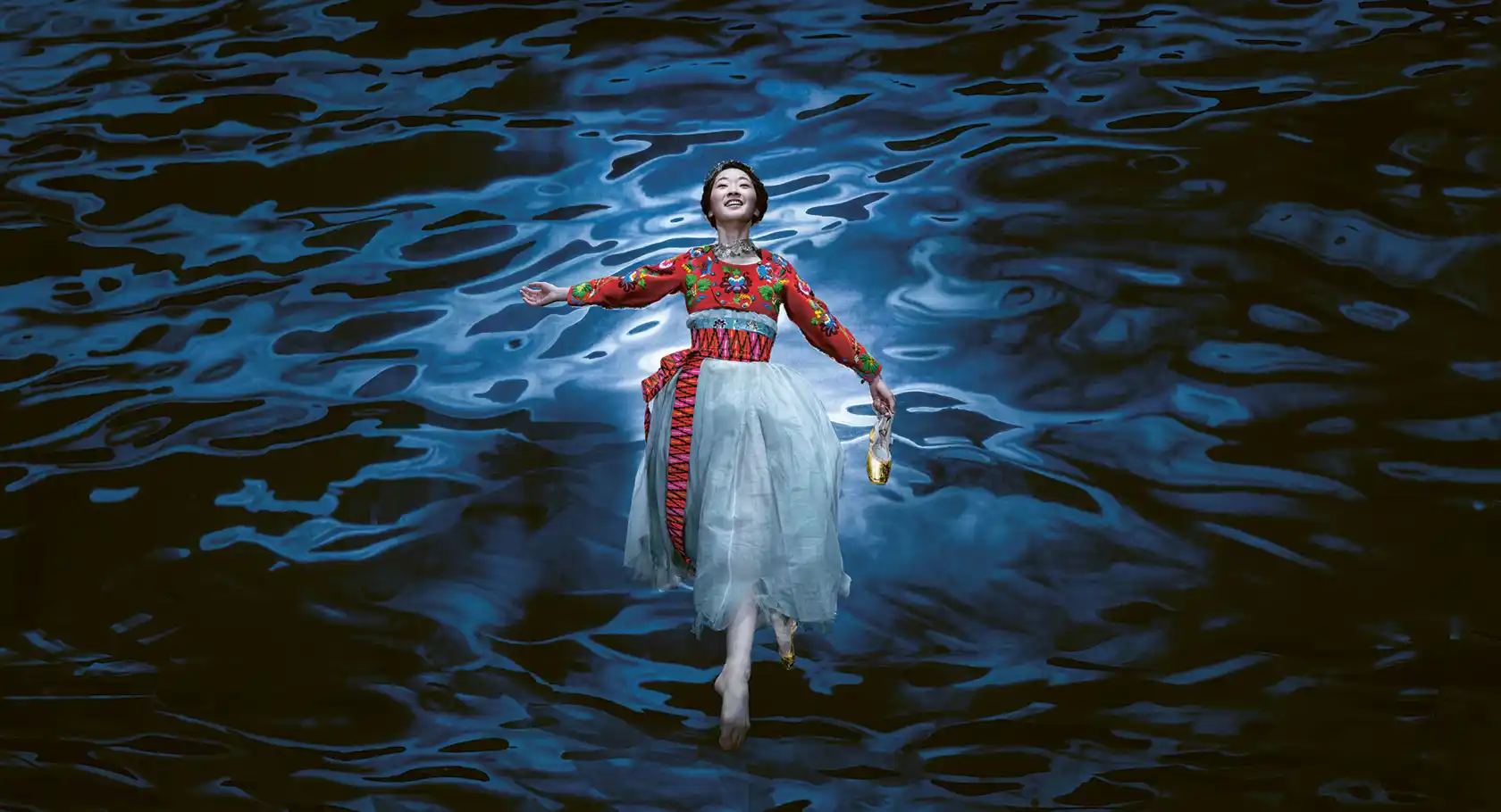 A woman, wearing a tulle skirt and folkloric jacket, floats over blue water: in her hand she holds a golden shoe.