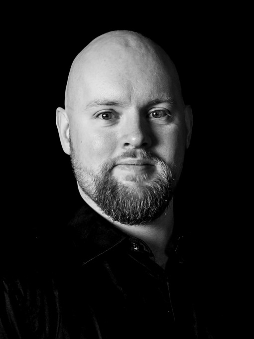 Närbild på en ung skäggig man som bär mörk kostym och mörk slips.