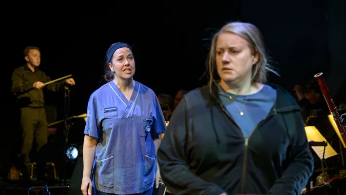 A woman in hospital clothes stands diagonally behind another woman, in regular clothes.