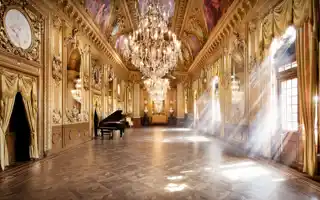 A gold-decorated room with a grand piano on the left, the sun shining through windows on the right.