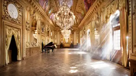 A gold-decorated room with a grand piano on the left, the sun shining through windows on the right.