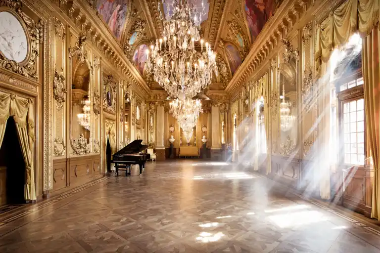 A gold-decorated room with a grand piano on the left, the sun shining through windows on the right.