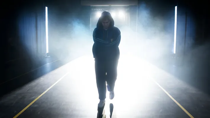 En pojke med armarna i kors och hängande blick står vänd mot kameran med skarpt motljus i bakgrunden.