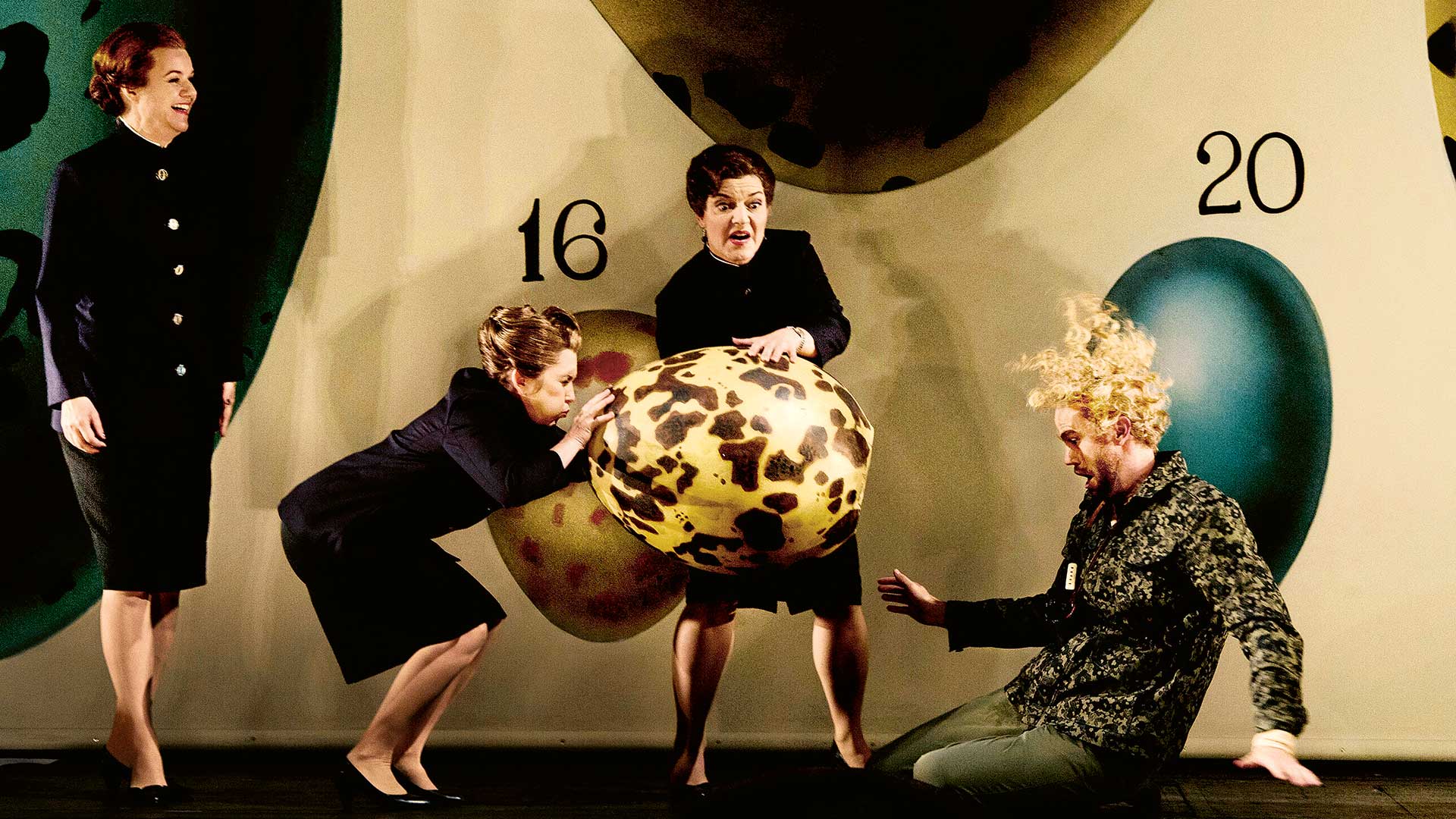 Två kvinnor håller i ett stort ägg, en man med lockigt hår sitter på golvet.