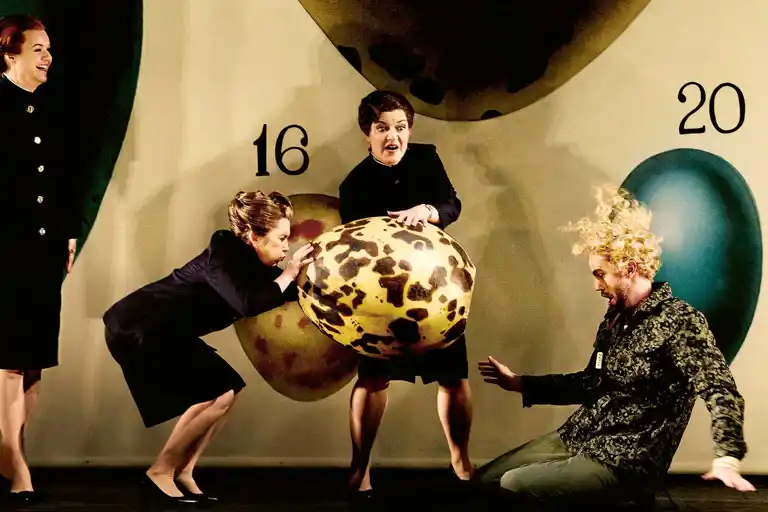 Två kvinnor håller i ett stort ägg, en man med lockigt hår sitter på golvet.