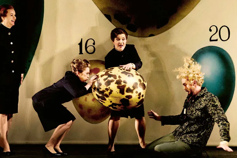 Två kvinnor håller i ett stort ägg, en man med lockigt hår sitter på golvet.