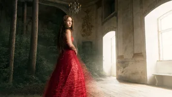 En kvinna i röd klänning står i ett rum med fönster, i vänstra hörnet växer en skog.