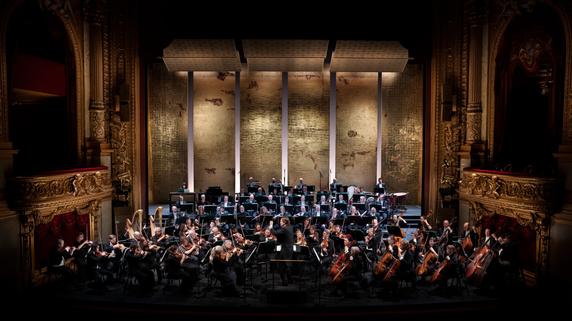 En stor orkester spelar på en scen som är omgärdad av gulddekorationer - i fonden en guldig kuliss.