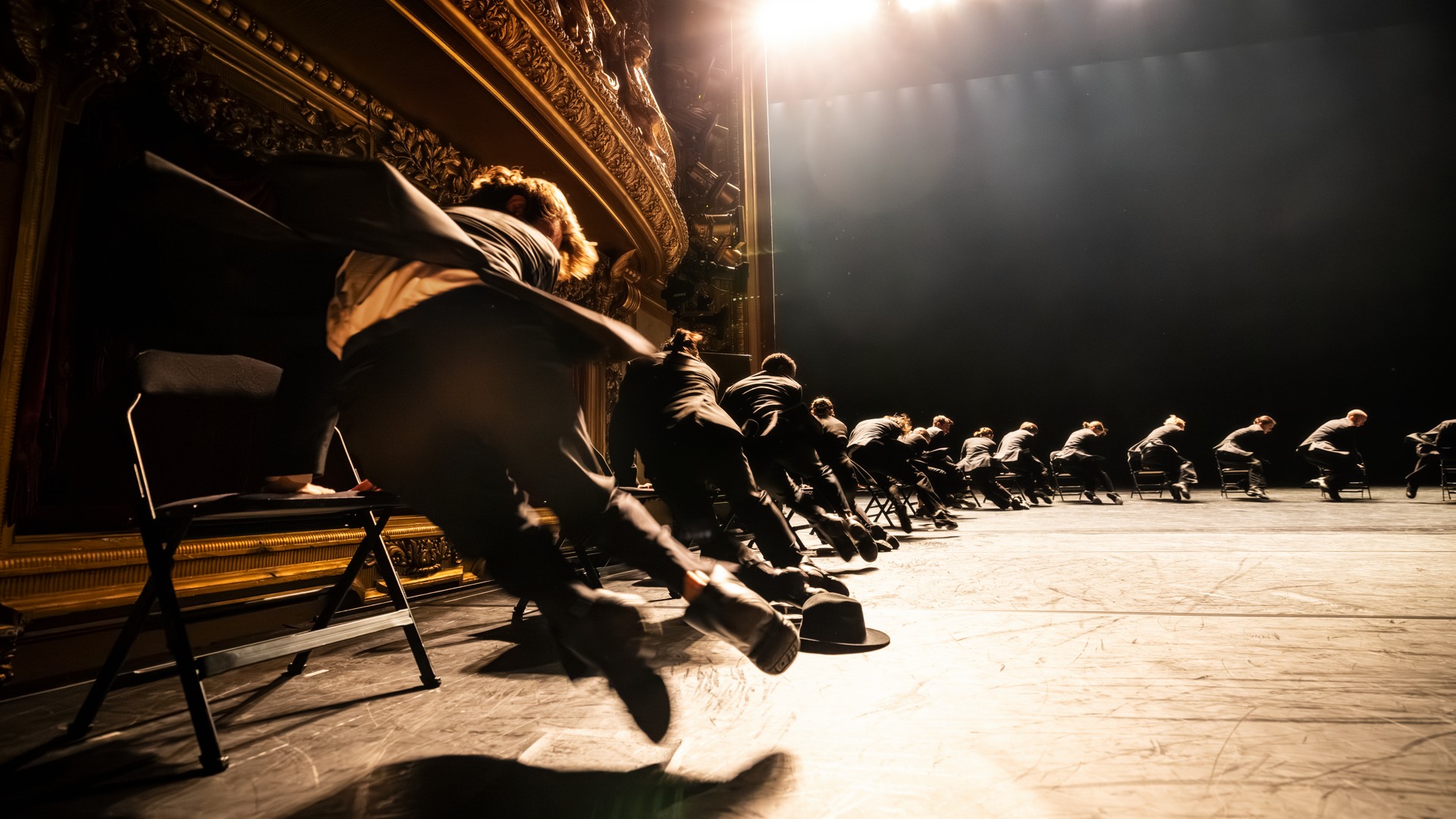 En mängd kostymklädda dansare i en halvcirkel, dansandes med stolar som stöd.
