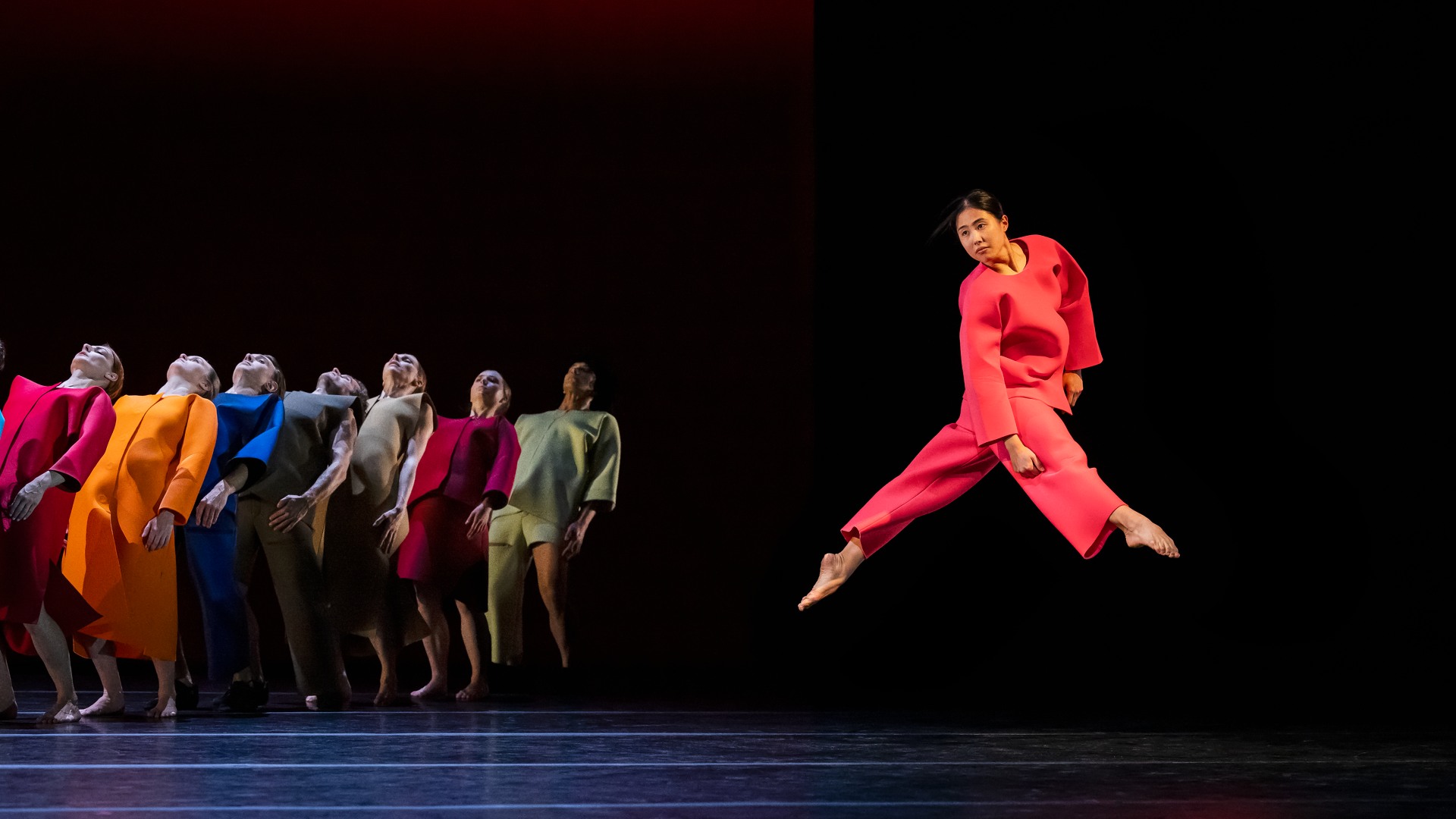 A group of dancers in colorful clothes, in the foreground a dancer in red jumps.